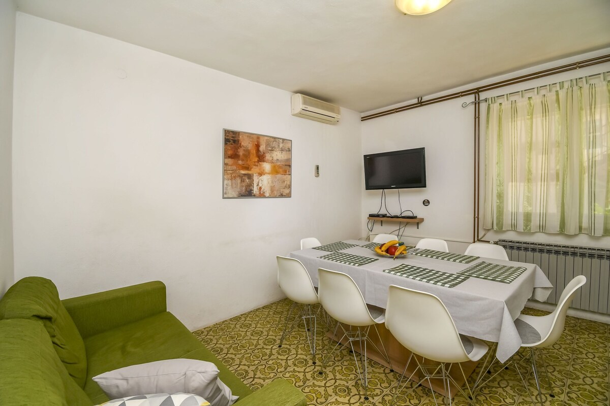 A room with a green sofa, a dining table with white chairs, a flat-screen TV, and a radiator.
