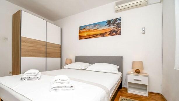 A neatly made bed with white linens and towels, a grey headboard, and a small bedside table with a lamp. - Apartment Zeljko (Makarska)