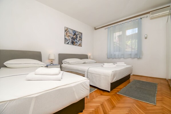 A hotel room with two beds, a wooden floor, a painting on the wall, and a window with a curtain. - Apartment Zeljko (Makarska)