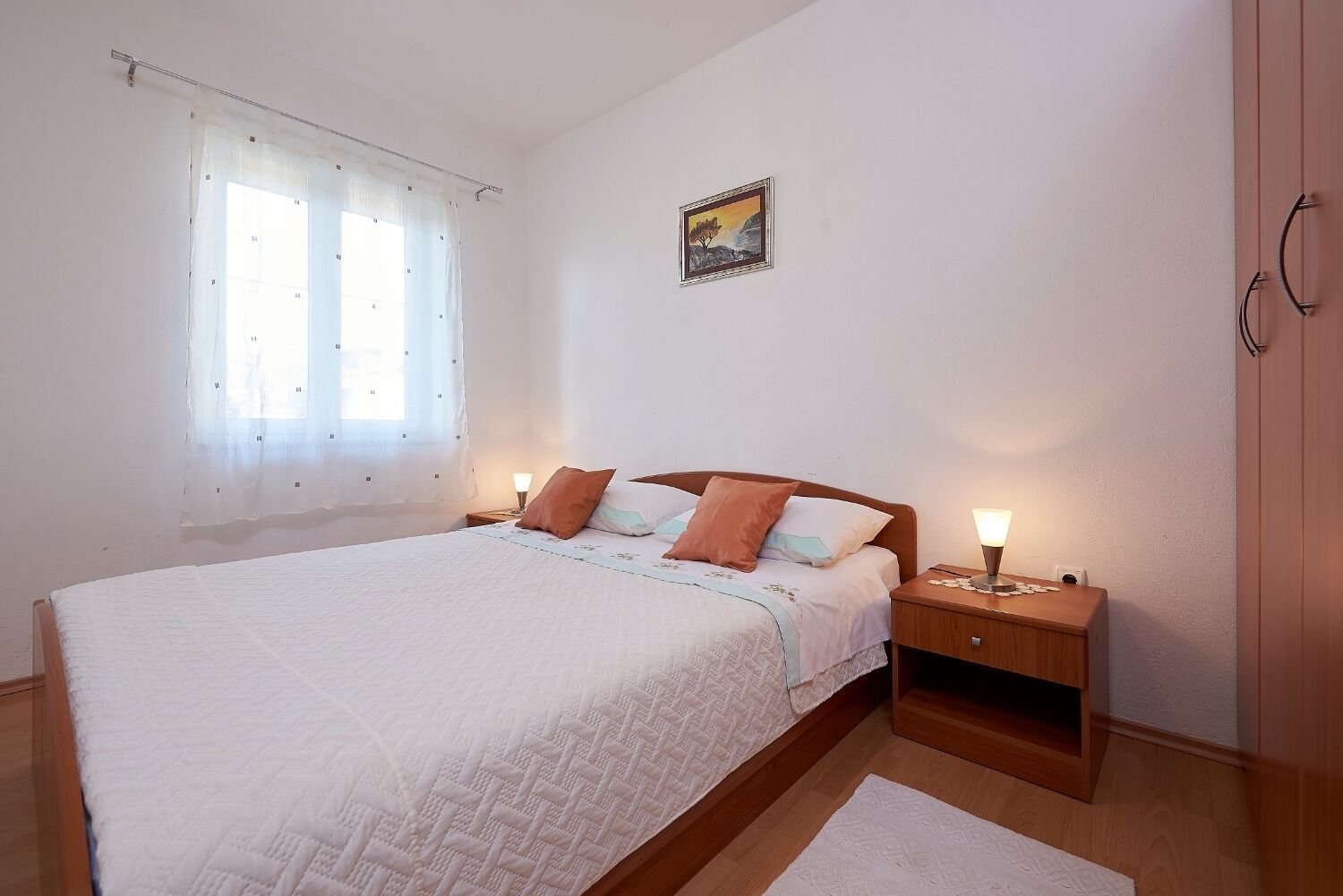 Une chambre à coucher comprenant un lit, des tables de chevet, une fenêtre avec des rideaux légers, un tableau au mur et une armoire en bois.