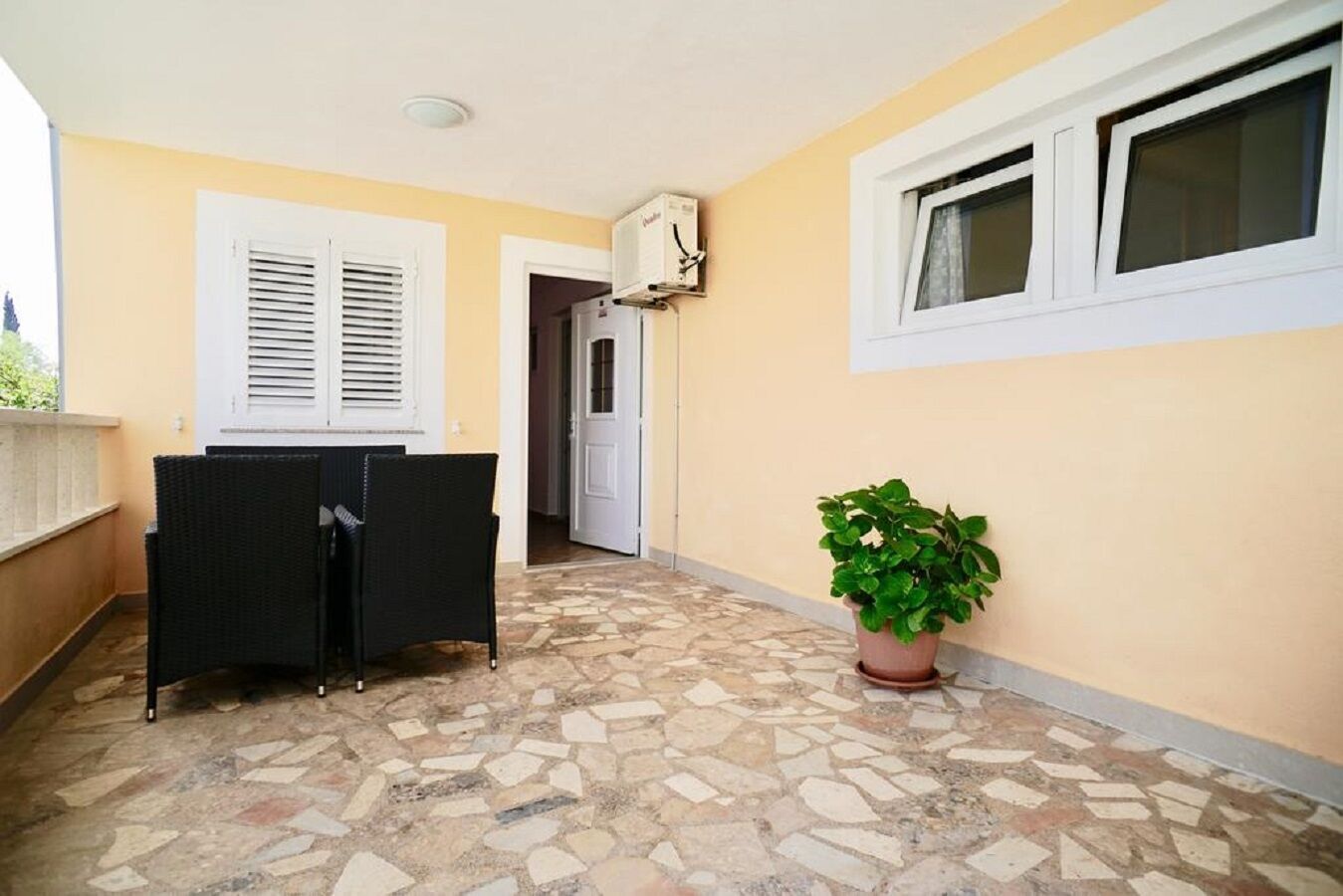 Un couloir avec un sol carrelé, une plante en pot, une porte et une fenêtre avec des stores.