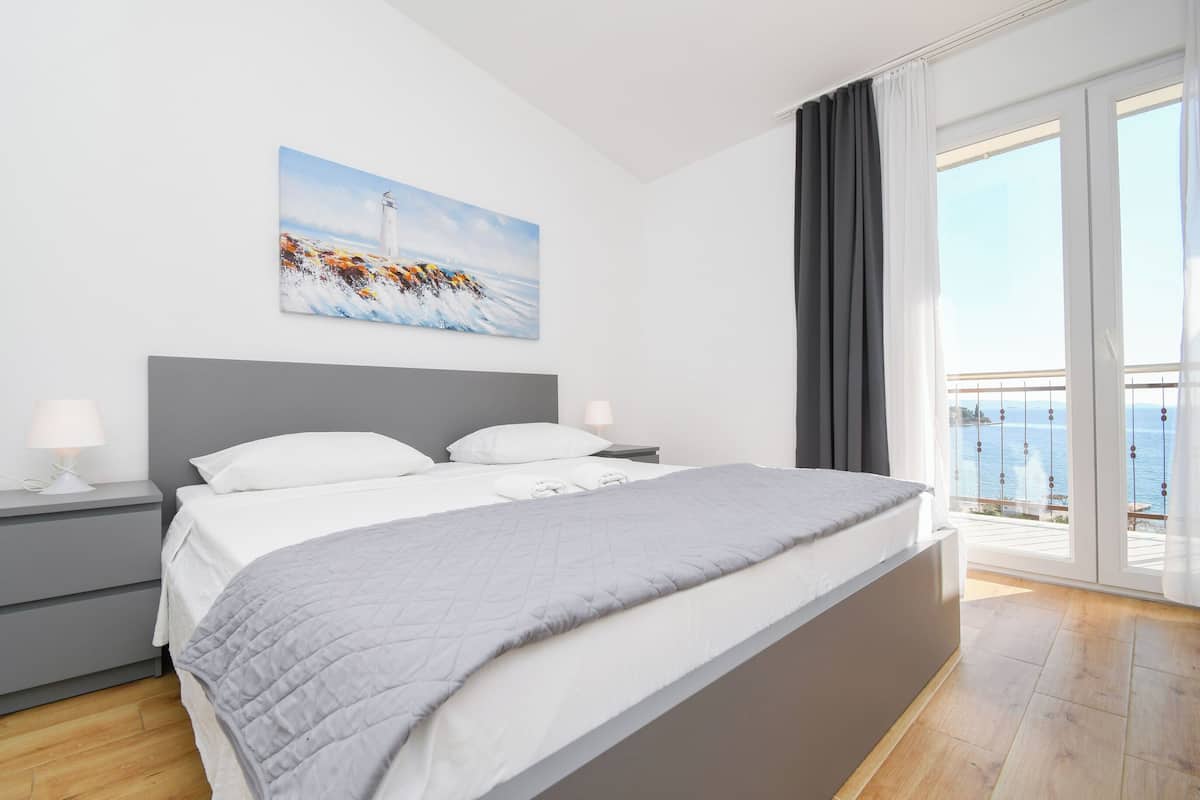 A bedroom with a large bed, a nightstand, a lamp, a painting of a lighthouse, and a balcony with a view of the sea.