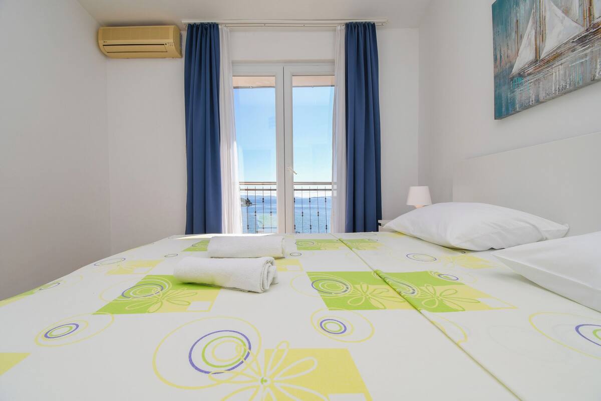 A hotel room with a bed, a window with curtains, a painting on the wall, and an air conditioning unit.