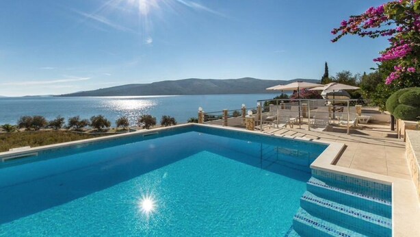 A clear blue swimming pool with steps leading into it, surrounded by outdoor seating and a view of the sea and mountains. - Apartments Rose (Seget)
