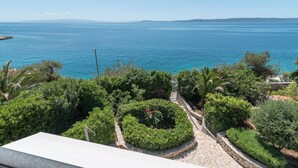 A view of a coastal area with a paved pathway leading to a circular garden with a fountain, surrounded by lush greenery and overlooking a calm sea. - Apartment Daniela (Okrug)