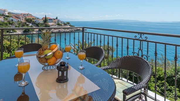 A round table with a fruit bowl, two glasses of orange juice, and a candle on a balcony overlooking the sea. - Apartment Daniela (Okrug)