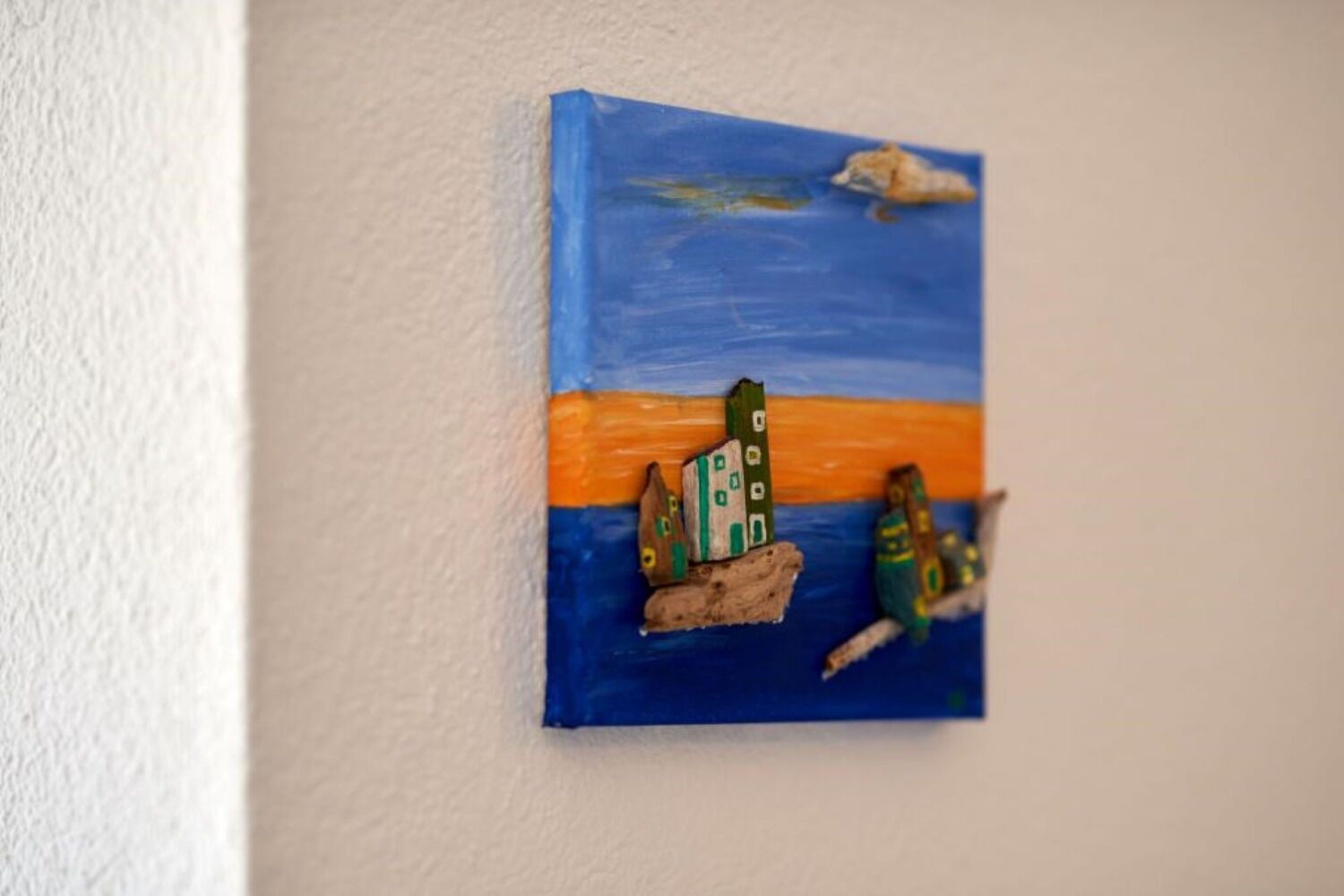 Een klein, kleurrijk schilderij van een boot met gebouwen, tegen een blauwe achtergrond, bevestigd aan een muur.