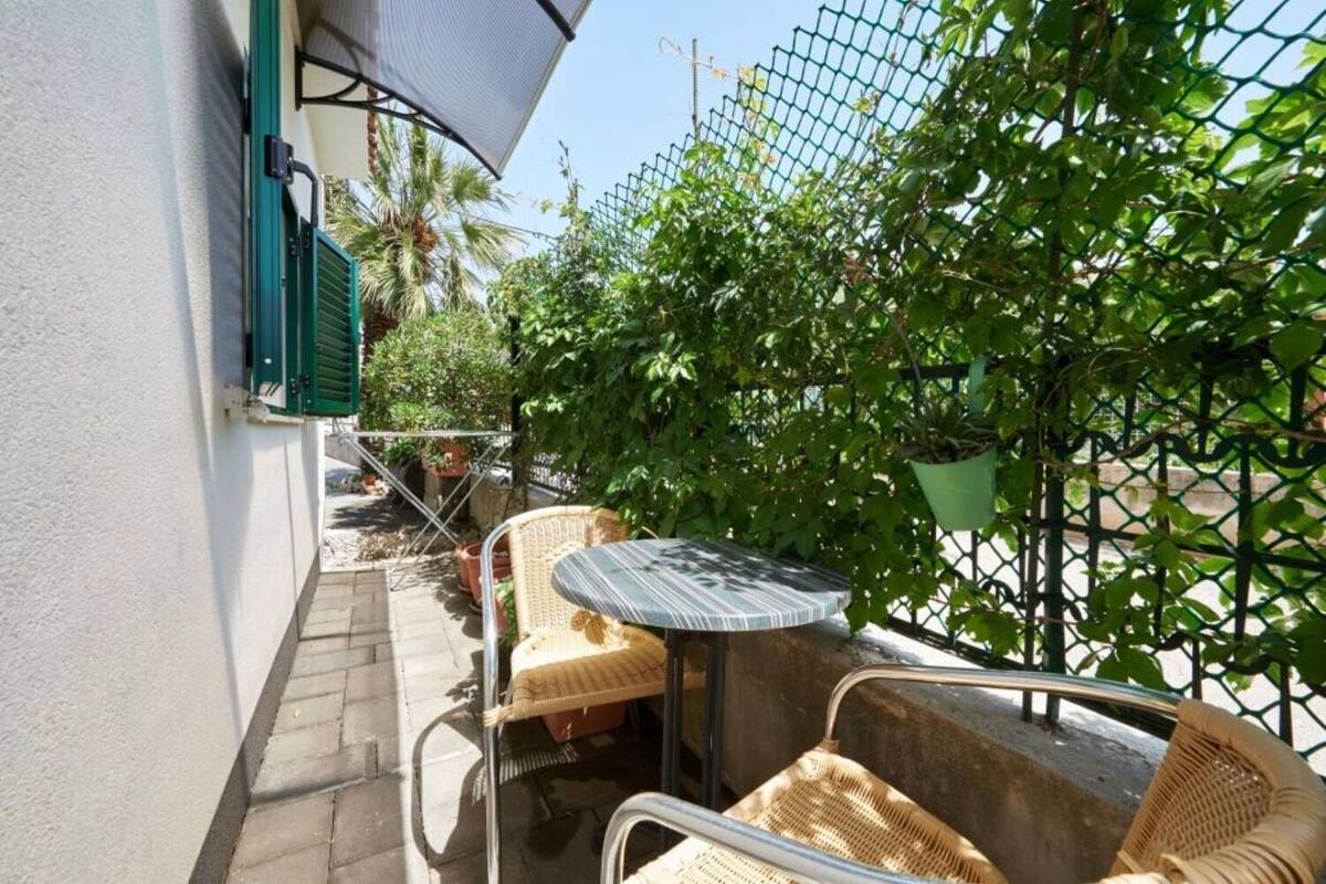 A small outdoor patio with a table and wicker chairs, surrounded by a green fence and plants.