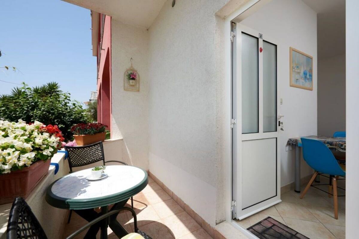 A small balcony with a table and chairs, a potted plant, and a door leading to a room with a bed and a chair.