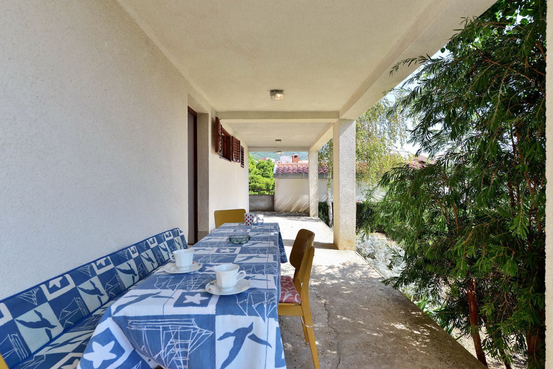 A covered outdoor dining area with a table set for two, a bench, and a view of the surrounding greenery and buildings.