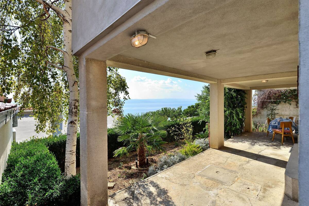A covered outdoor area with a view of the sea, a table with chairs, and various plants.