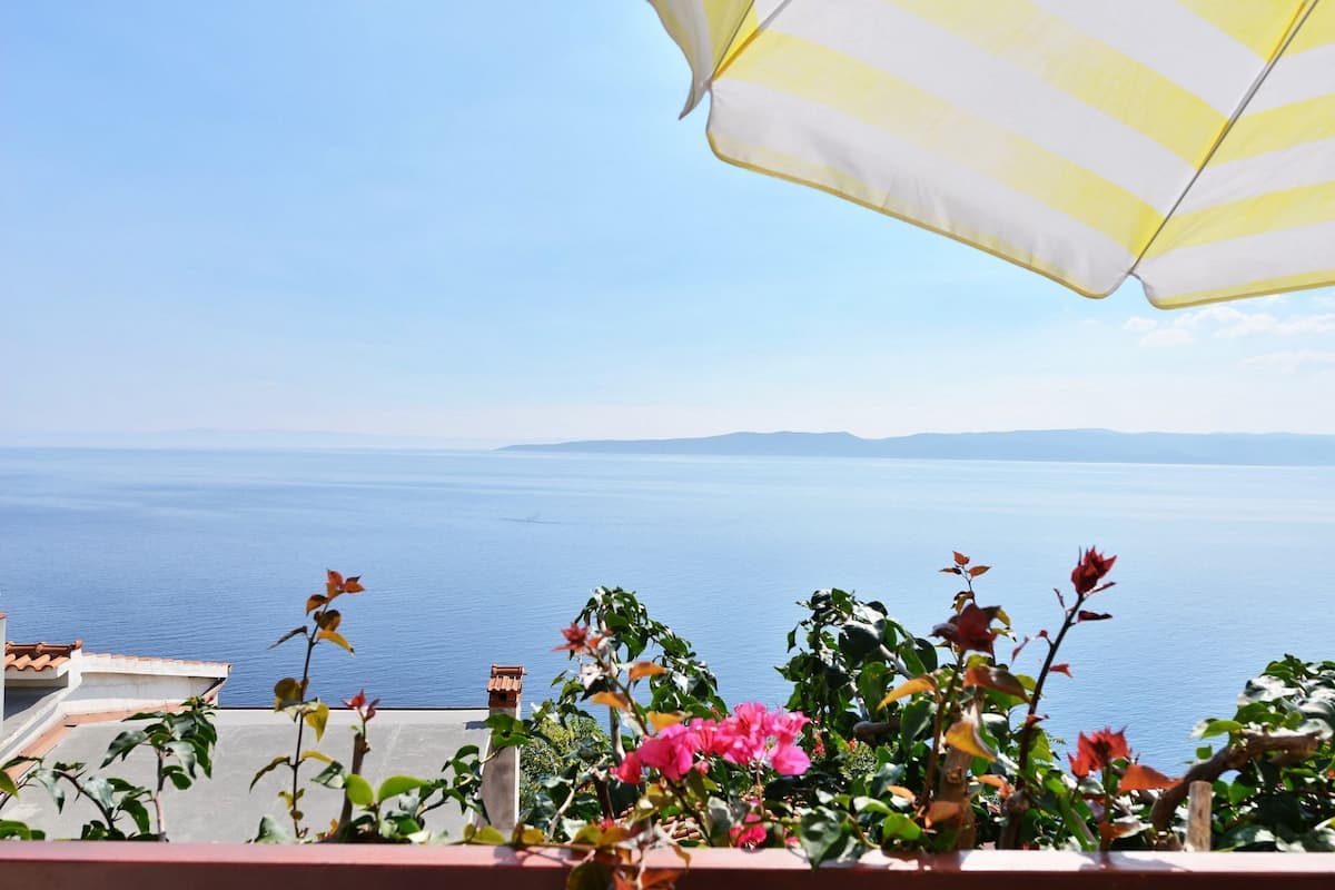 A balcony with a view of the sea, a striped umbrella, and potted plants.