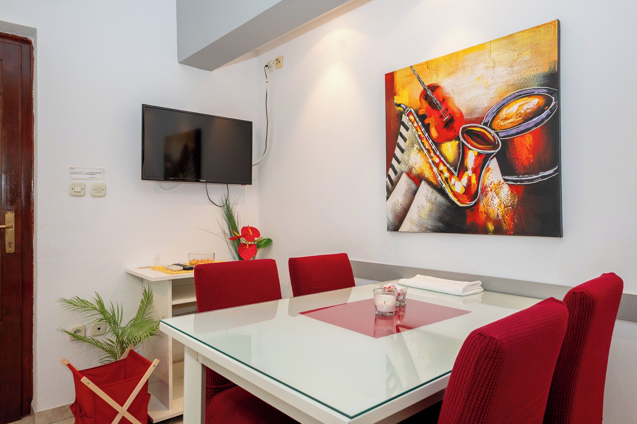 Un moderno comedor con mesa de vidrio, sillas rojas, televisor de pantalla plana en la pared y un cuadro abstracto lleno de color.
