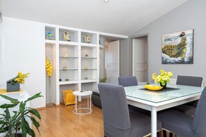 A modern dining area with a glass table, grey upholstered chairs, and a white shelving unit with decorative items. - Apartments Antisa (Tucepi)