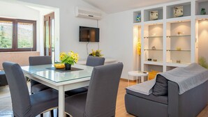 A modern living room with a glass dining table, grey upholstered chairs, a grey sofa, a flat-screen TV mounted on the wall, and a built-in shelving unit with decorative items. - Apartments Antisa (Tucepi)