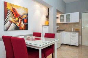 A modern kitchen with white cabinets, a glass dining table with red chairs, and a painting of musical instruments on the wall. - Apartments Antisa (Tucepi)