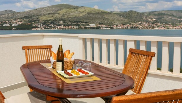 A wooden table with a bottle of wine, bread, and a plate of olives on a balcony overlooking a coastal town and hills. - House Dupla (Okrug)