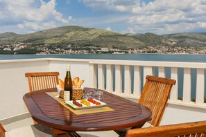 A wooden table with a bottle of wine, bread, and a plate of olives on a balcony overlooking a coastal town and hills. - House Dupla (Okrug)