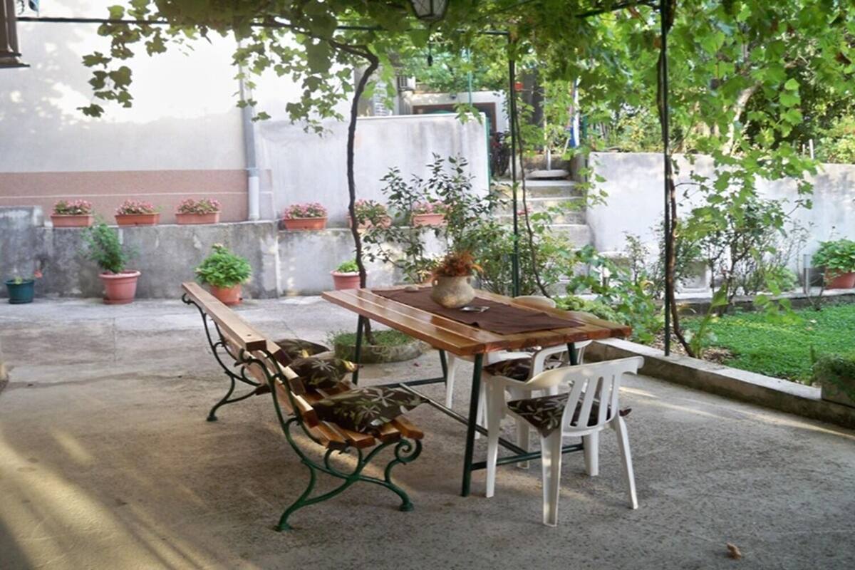 An outdoor patio with a wooden table and benches, surrounded by potted plants and a concrete floor.