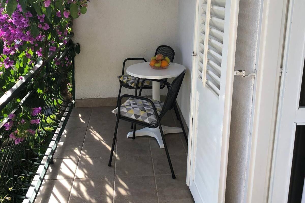 A small outdoor seating area with a table and chairs, a basket of fruit, and a wall with a window and a door.