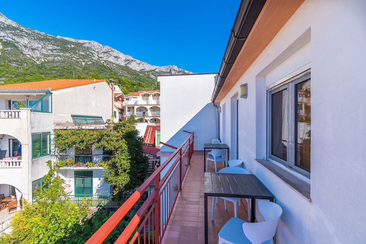 A balcony with a table and chairs, overlooking a mountainous area and neighboring buildings.