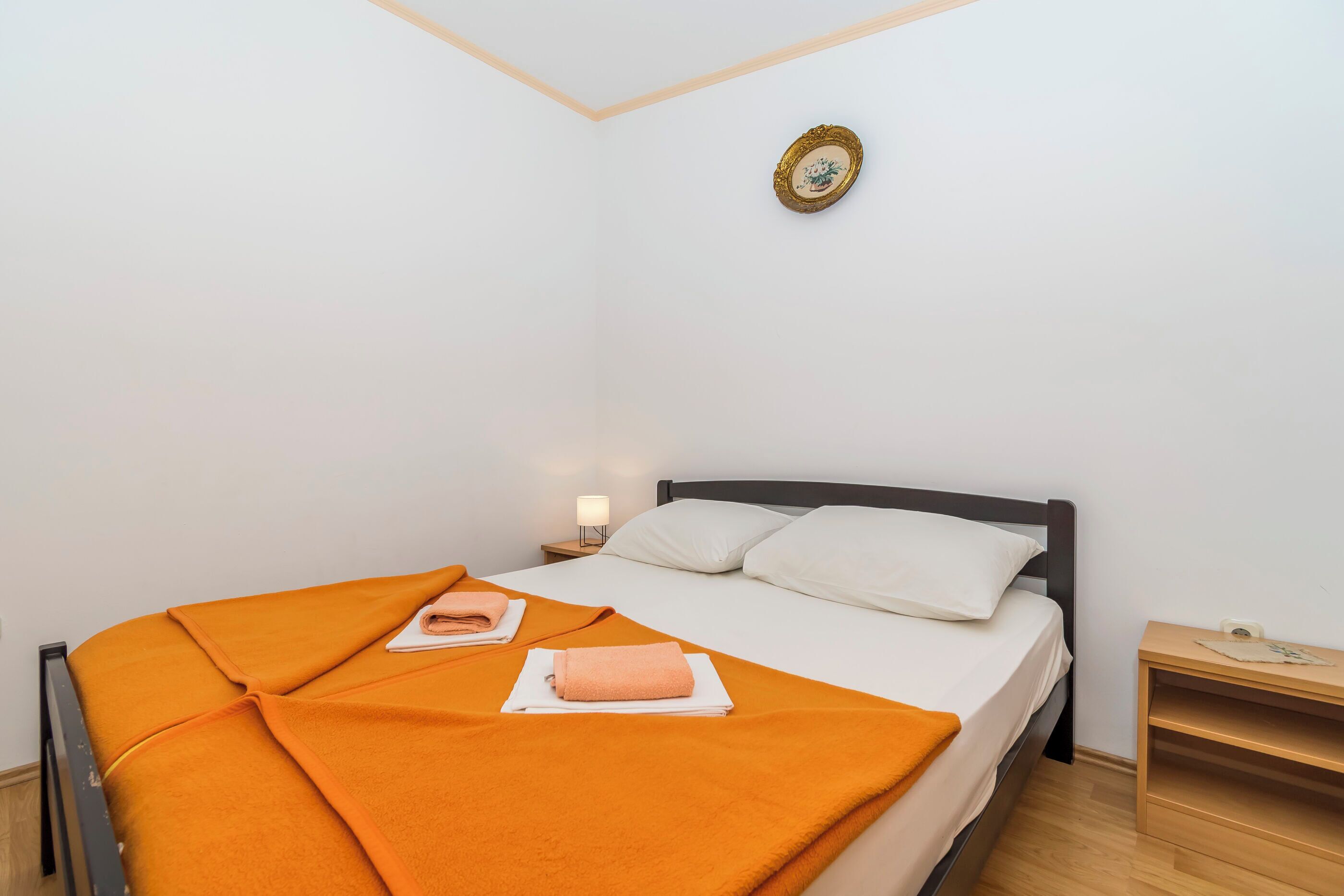 Ein ordentlich bezogenes Bett mit einer orangefarbenen Decke, ein Nachttisch mit einer Lampe und ein gerahmtes Bild an der Wand.