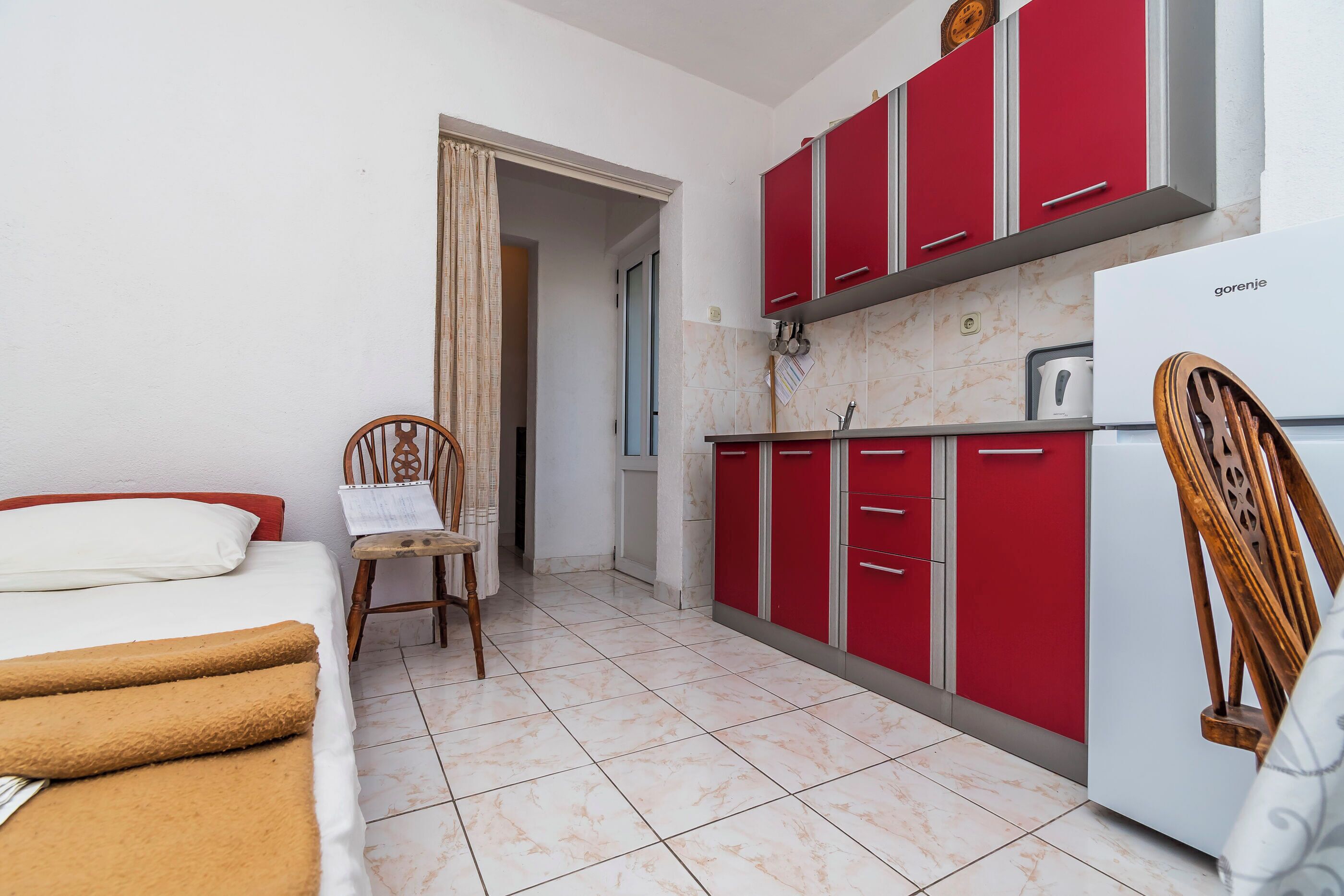 A compact room with a bed, a chair, a kitchenette with red cabinets, and a white refrigerator.
