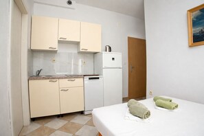A compact hotel room with a kitchenette, a bed with two green towels, and a wooden door. - Apartments Tiho (Podgora)