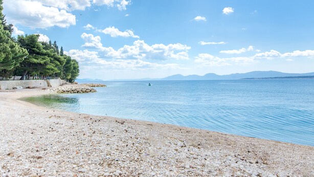 A pebble beach with a calm sea, clear sky, and distant mountains. - Apartments Tiho (Podgora)