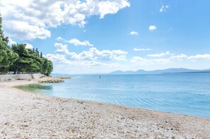 A pebble beach with a calm sea, clear sky, and distant mountains. - Apartments Tiho (Podgora)