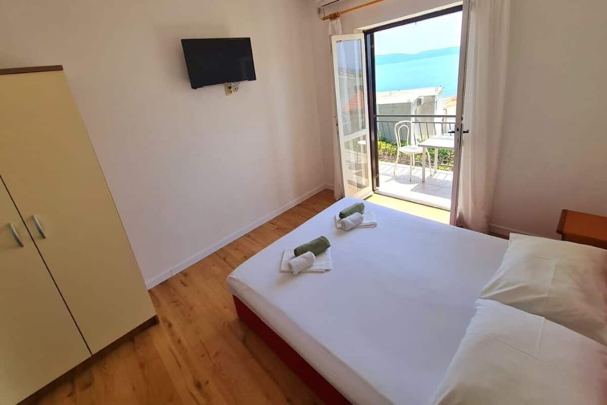 A hotel room with a bed, a television mounted on the wall, and a sliding glass door leading to a balcony with a view of the sea.