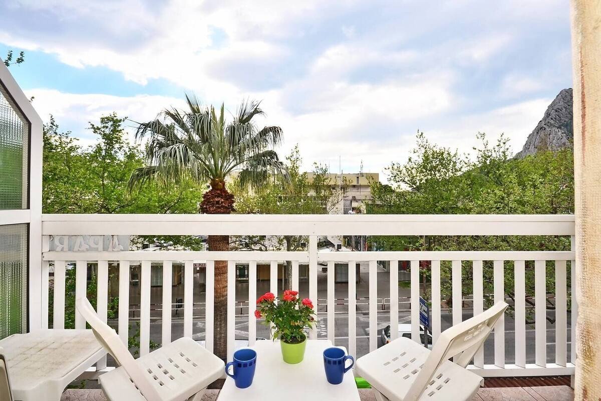 A balcony with white railings, a table with two chairs, a potted plant with red flowers, and a view of greenery and a mountain in the background.