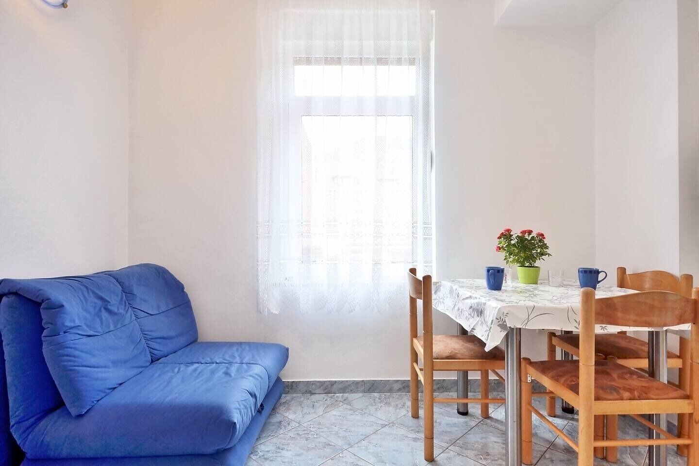 A room with a blue sofa, a dining table with chairs, a window with sheer curtains, and a potted plant on the table.