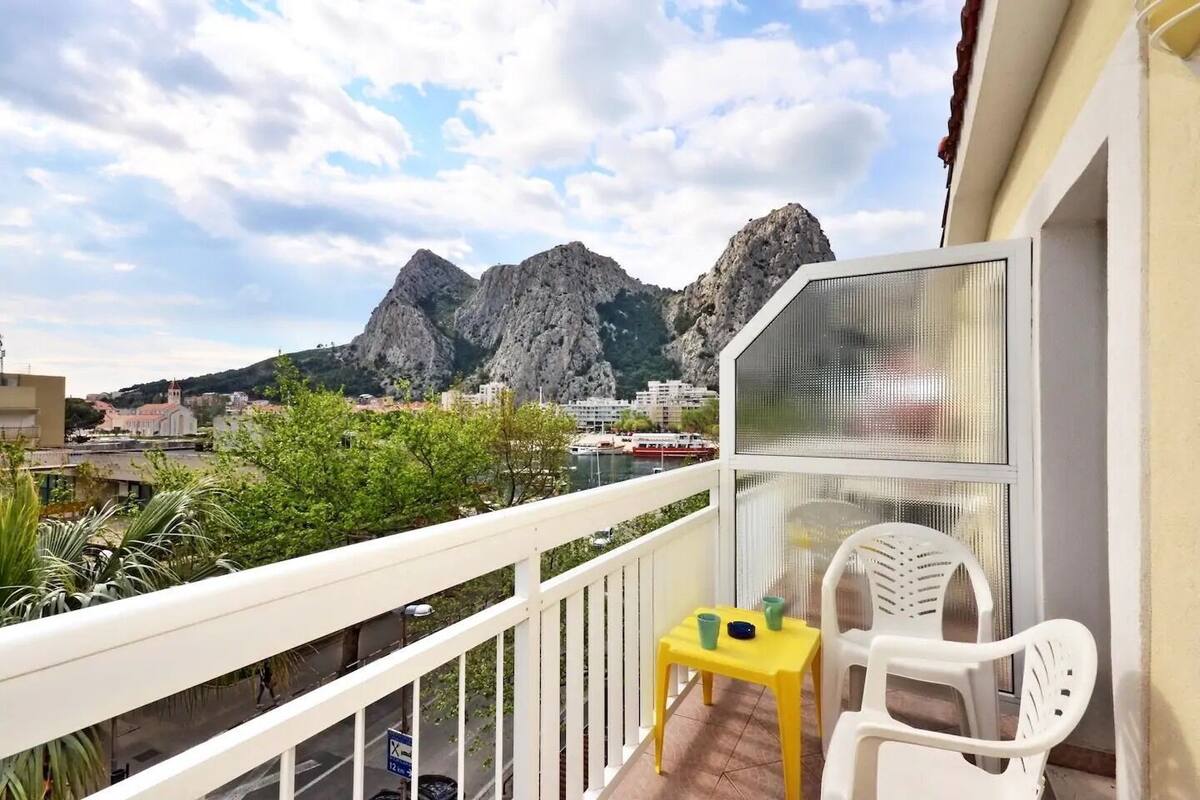 A balcony with a yellow table and white chairs, overlooking a mountainous landscape and a body of water with boats.
