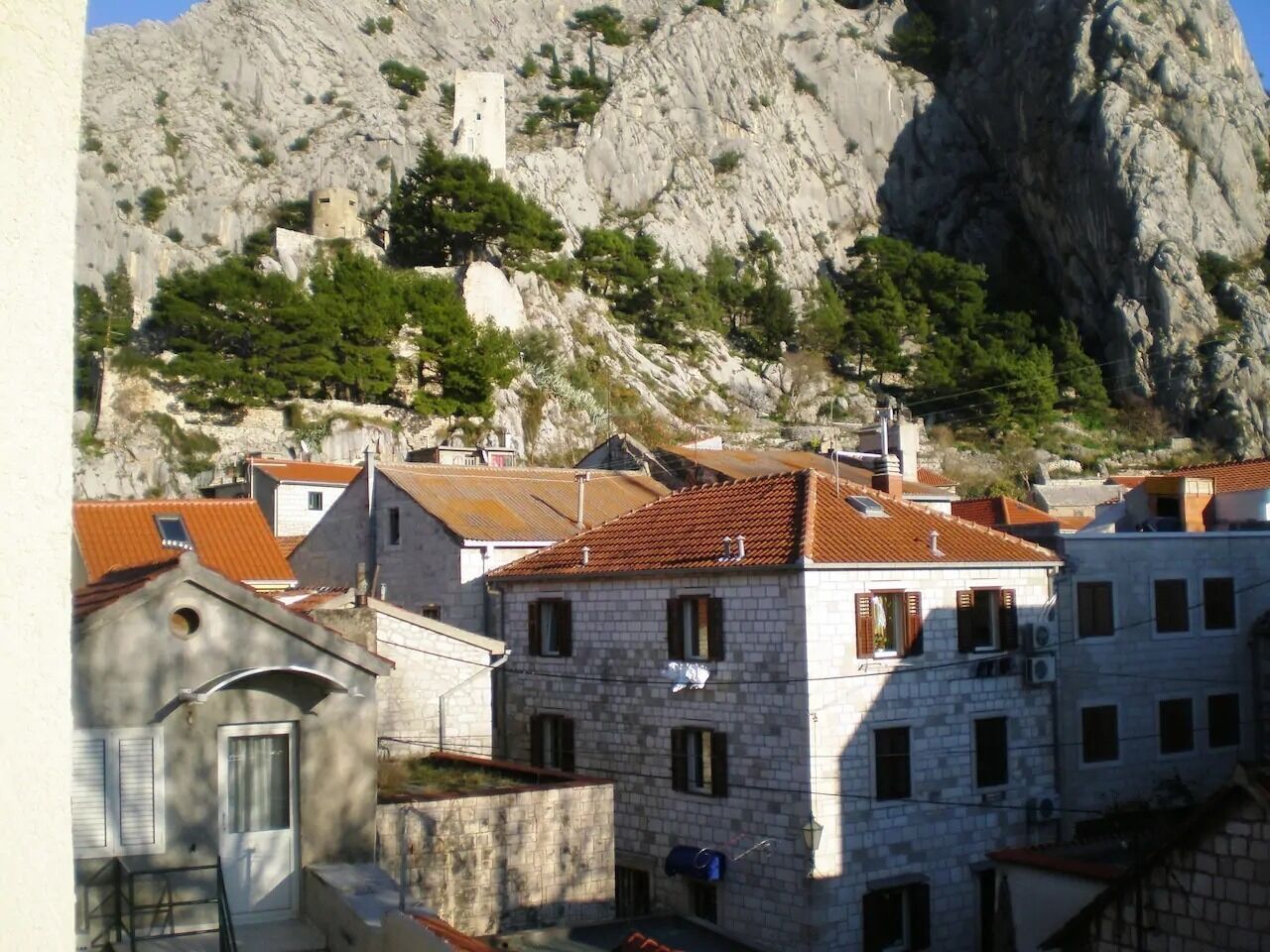 Un edificio de piedra con techo de tejas rojas, rodeado de otros edificios y un promontorio rocoso.