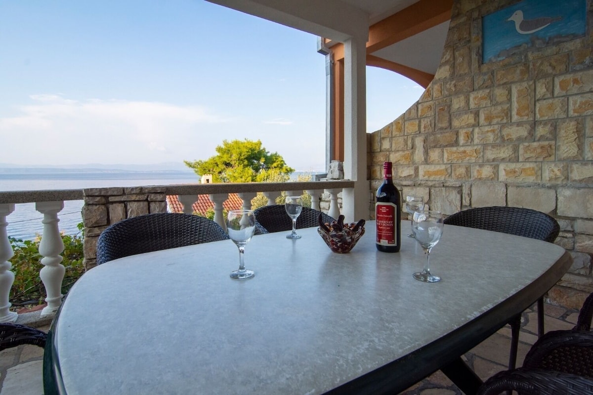 A table set for two with a bottle of wine, glasses, and a decorative bowl on a balcony overlooking a body of water.