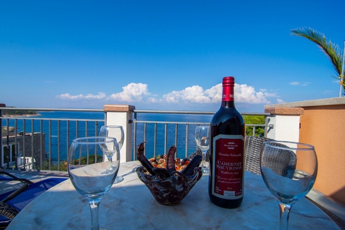 A table with a bottle of Croatian wine, two glasses, and a decorative bowl on a balcony overlooking the sea.