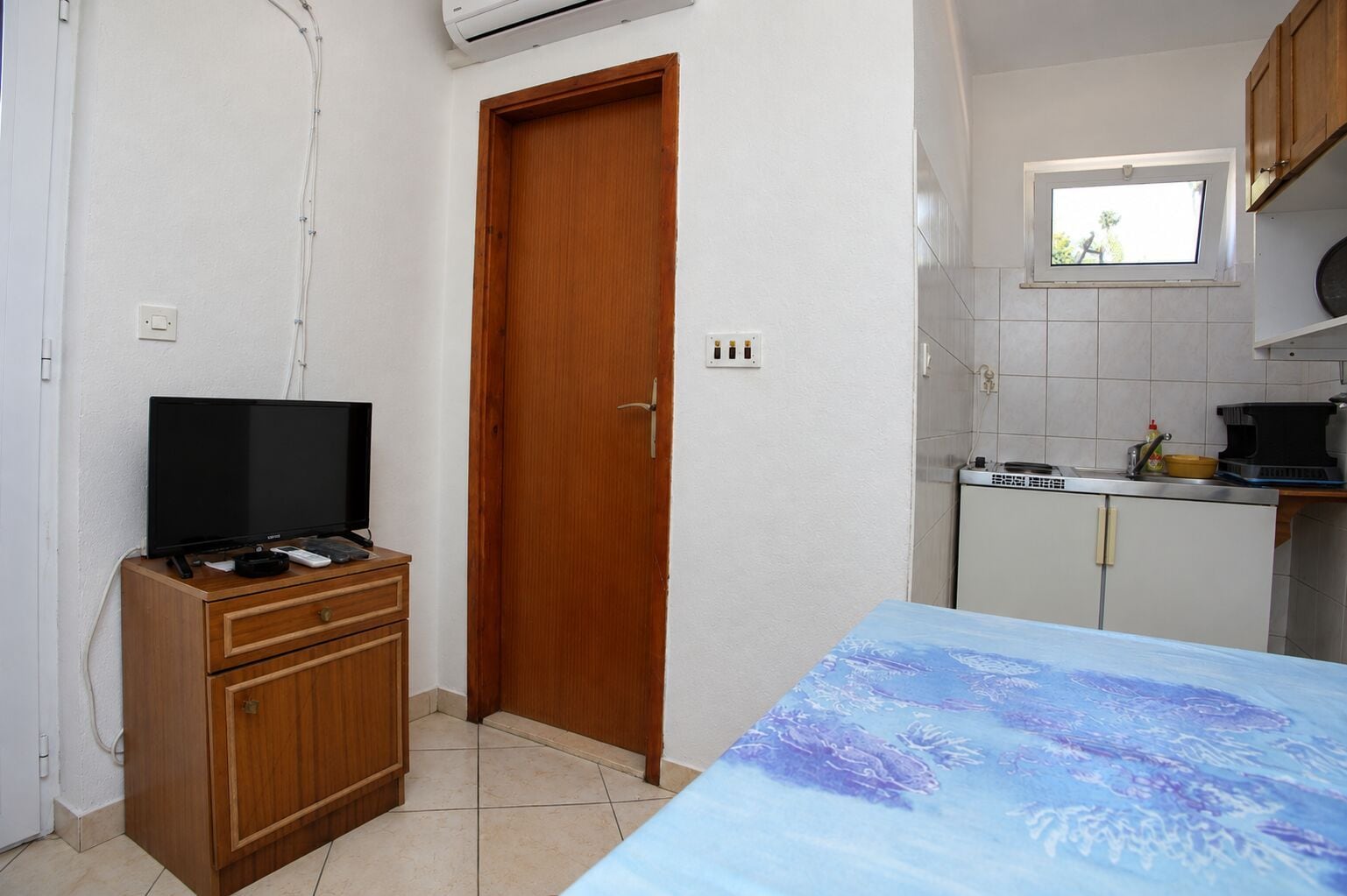 Une petite pièce comprenant un meuble en bois, une télévision, un lit recouvert d’une courtepointe à motifs bleus, une fenêtre et une kitchenette équipée d’une cuisinière et d’un évier.