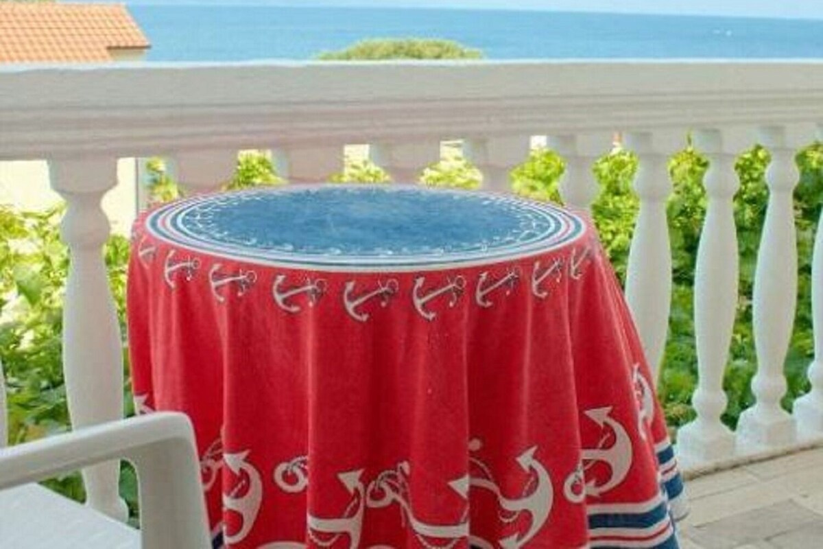 A round table with a red tablecloth featuring white anchor designs, set on a balcony with a white balustrade.