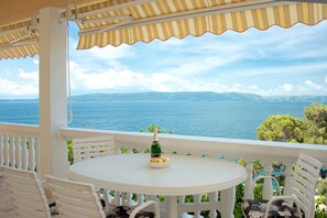A covered outdoor seating area with a table and chairs, overlooking a body of water and distant hills. - Apartment Gojko (Podgora)