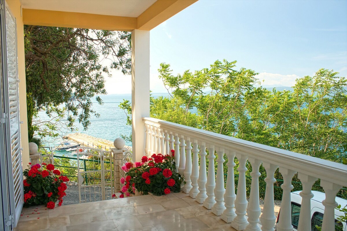 A balcony with a white balustrade, red flowers, and a view of the sea and greenery.