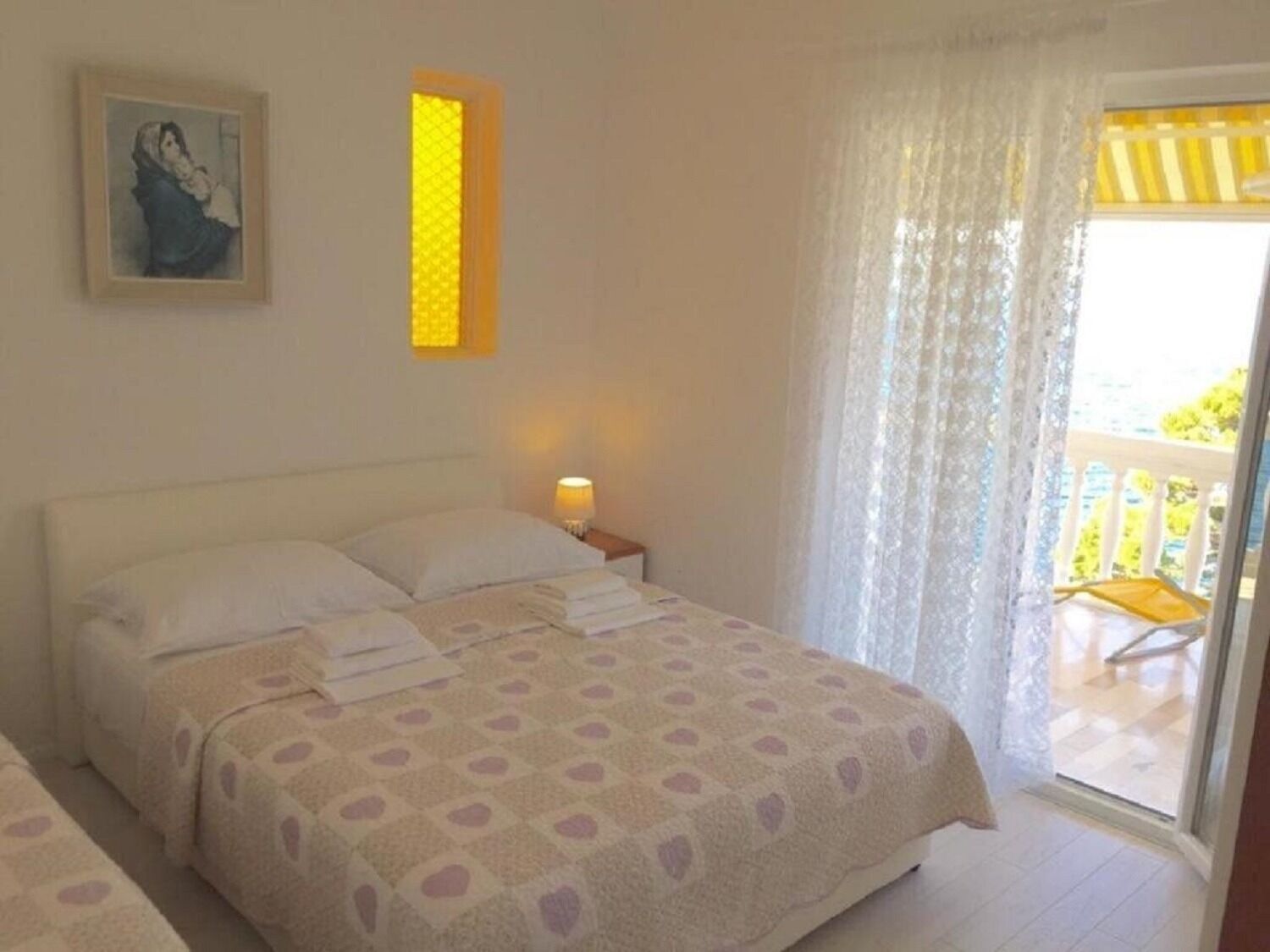 Quarto com cama, mesa de cabeceira, luminária, quadro e varanda com cadeira.