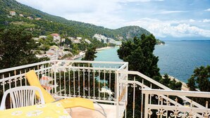 A balcony with a white metal railing overlooking a coastal town, a chair with a yellow cushion, and a view of the sea and hills. - Apartment Gojko (Podgora)