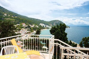A balcony with a white metal railing overlooking a coastal town, a chair with a yellow cushion, and a view of the sea and hills. - Apartment Gojko (Podgora)