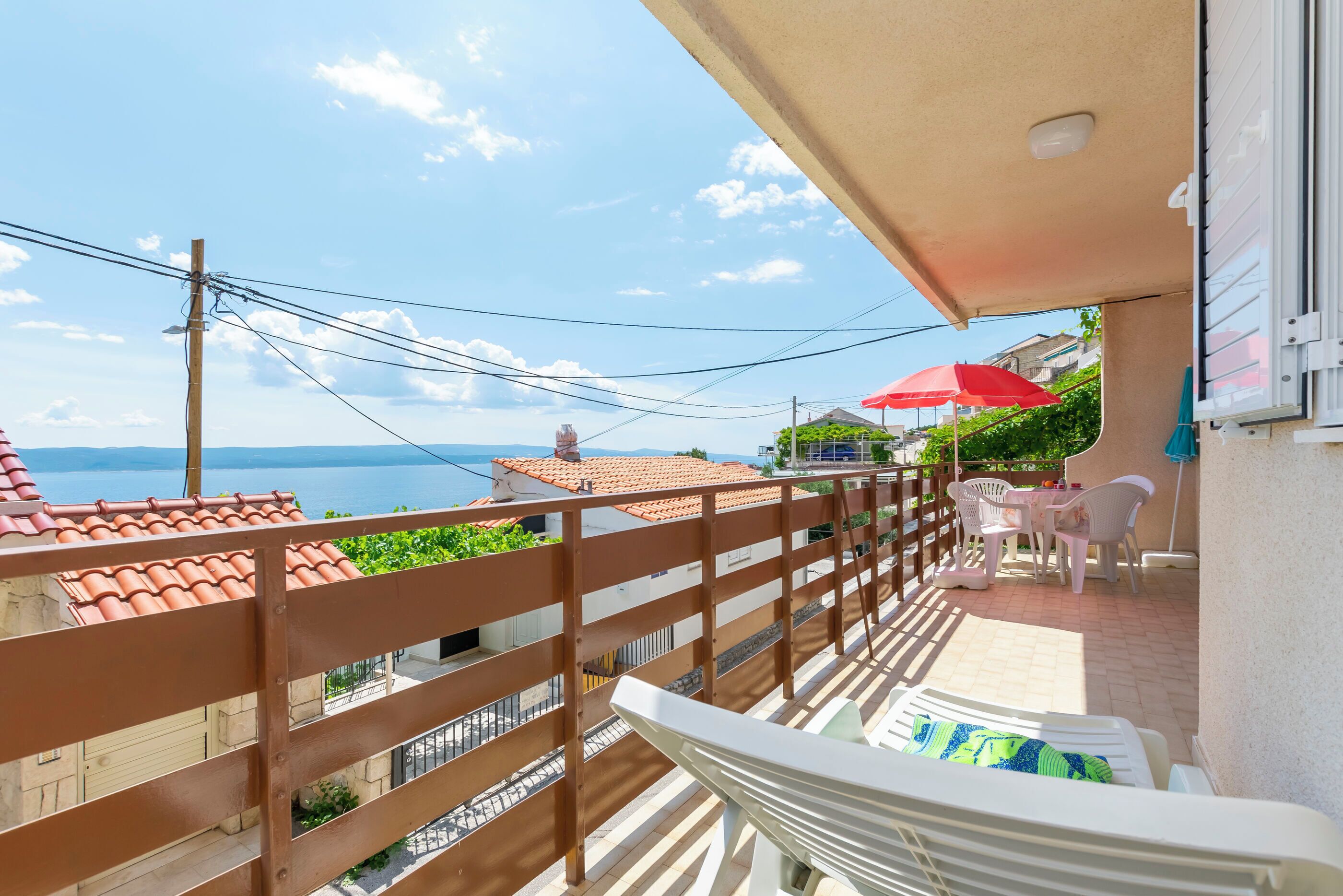 A balcony with a view of the sea, a red umbrella, and a table with chairs.
