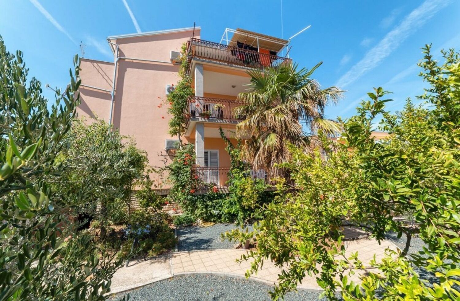 A multi-story building with balconies, surrounded by lush greenery and a clear sky.