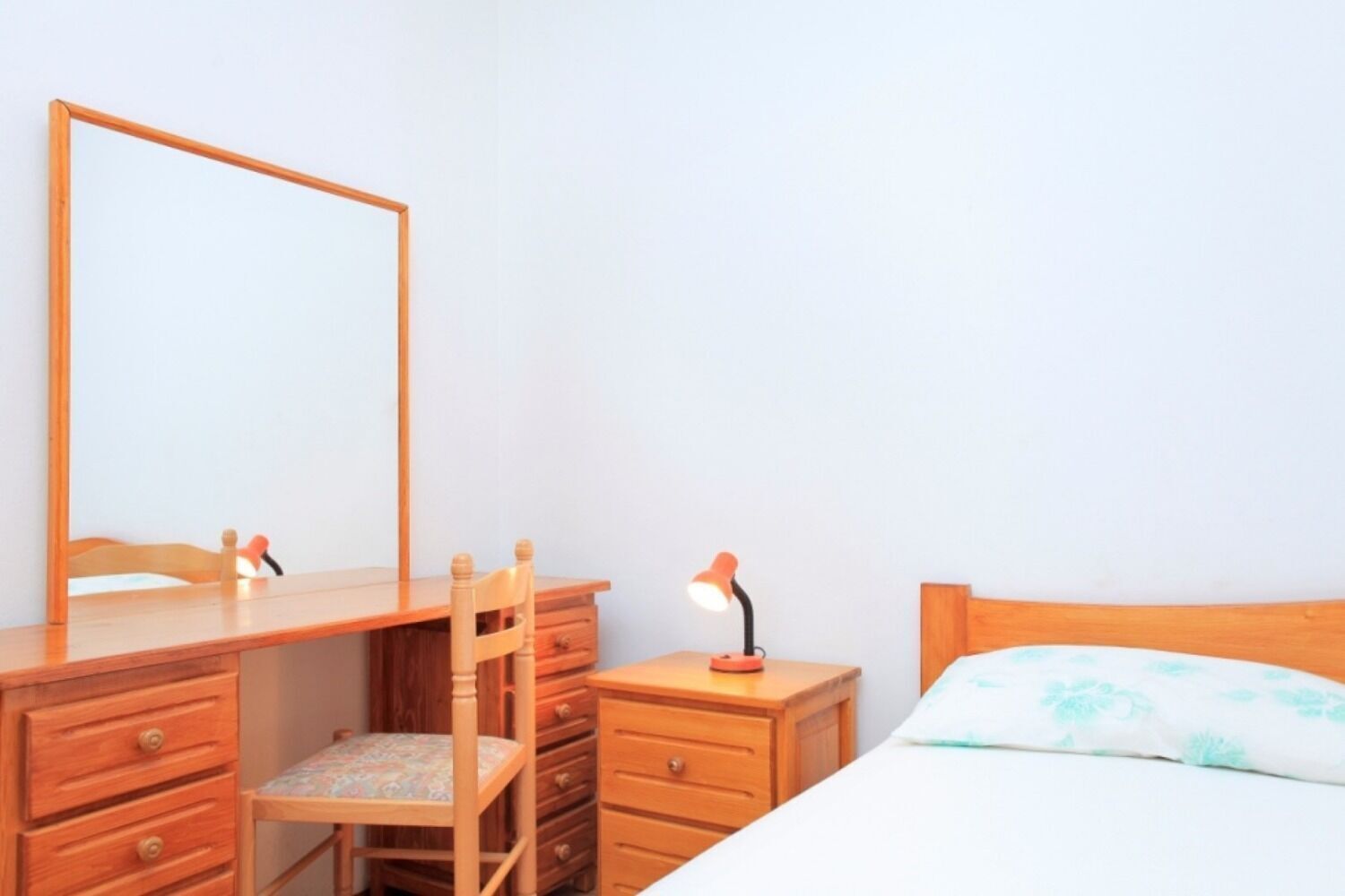 A bedroom with a wooden dresser, a chair, a bedside table with a lamp, and a bed with a floral bedspread.
