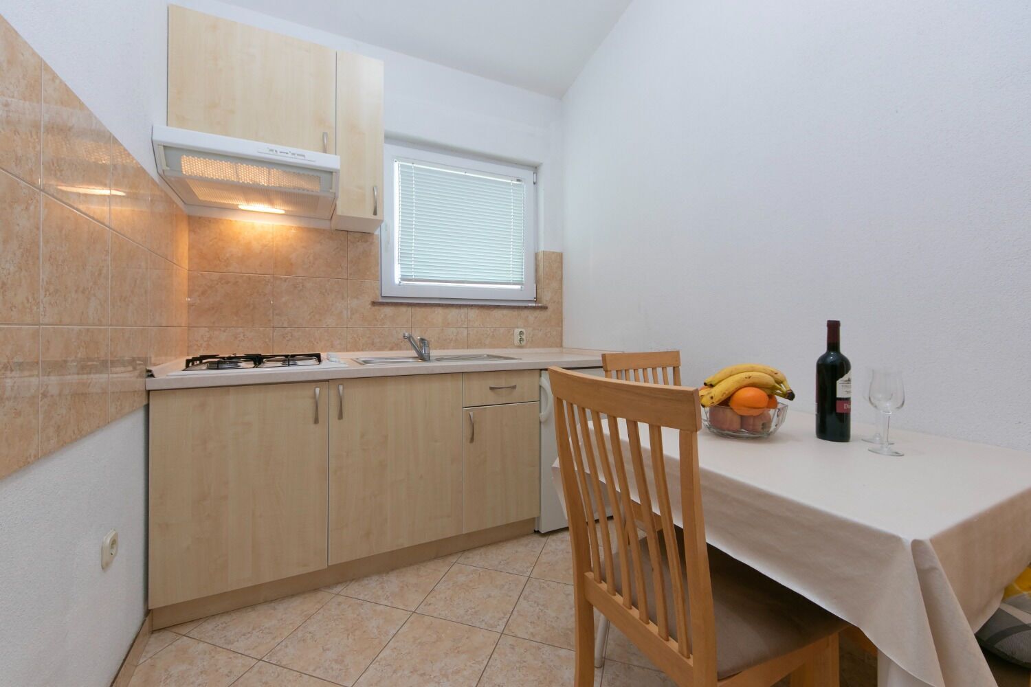 A compact kitchen with a gas stove, sink, and wooden cabinets. There's a window with blinds, a table with chairs, and a bowl of fruit and a bottle of wine on the table.