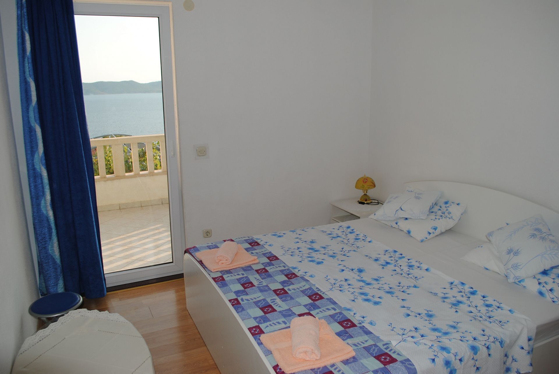 A bedroom with a bed, a nightstand, a lamp, and a view of the sea through a glass door.