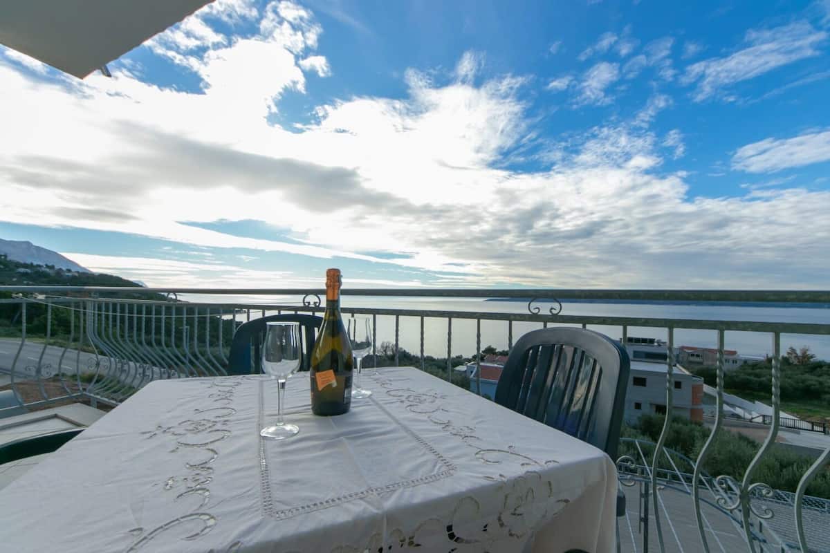 A balcony table with a bottle of wine and glasses, overlooking a body of water and buildings.